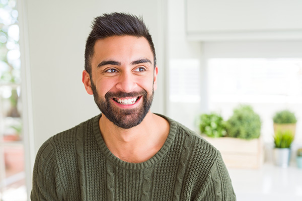 A smiling man with a beard, wearing a dark sweater, seated indoors near potted plants.