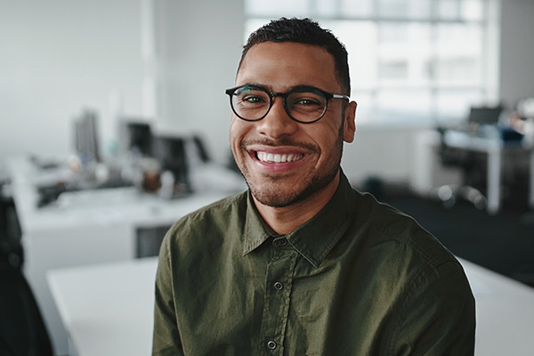The image shows a smiling man in glasses, wearing a green shirt and sitting at a desk in an office environment.