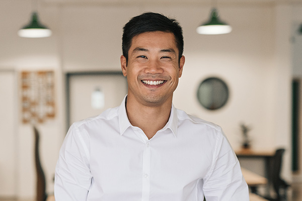 The image features a man with a smile, wearing a white shirt, standing in an interior setting with a modern office environment, against a white wall with framed items and a light fixture overhead.
