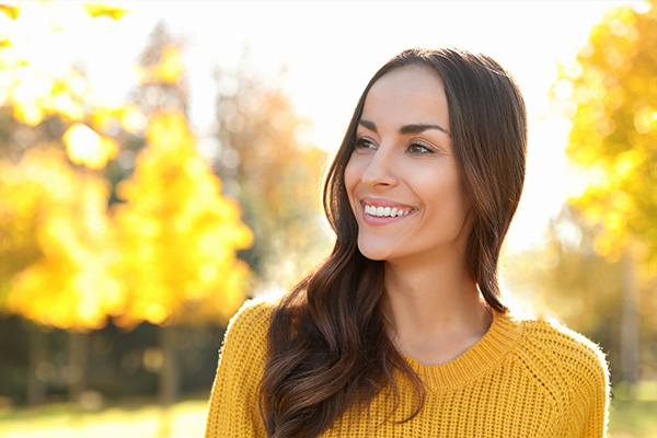 A smiling woman with long hair stands in a park during autumn, wearing a yellow sweater and standing against a blurred background of trees and foliage.