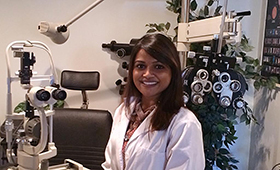 The image shows a woman standing in front of an eye examination machine, wearing a white lab coat, with a smile on her face.