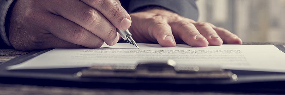 The image shows a person s hand holding a pen over a piece of paper on a desk, with a blurred background suggesting an office setting.