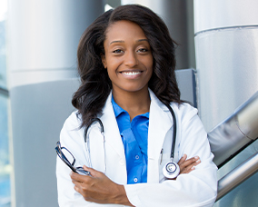 A woman wearing a white lab coat stands confidently with her arms crossed in front of a modern building entrance, smiling at the camera.