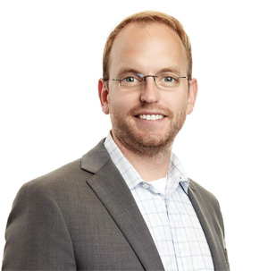 The image shows a man standing against a white background, wearing glasses, a light-colored shirt with a tie, and a dark suit jacket over a patterned shirt. He has short hair, a beard, and is smiling at the camera.