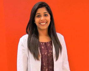 The image shows a woman standing against a red background, smiling at the camera. She has long hair, is wearing a white lab coat, and has a lanyard around her neck.